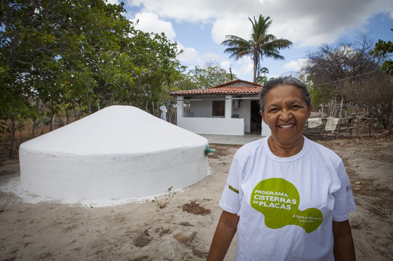 Agricultora negra usando blusa branca com o texto "Programa Cisternas de Placas". Ao lado dela uma cisterna recém pintada de branco se destaca à frente da casa.